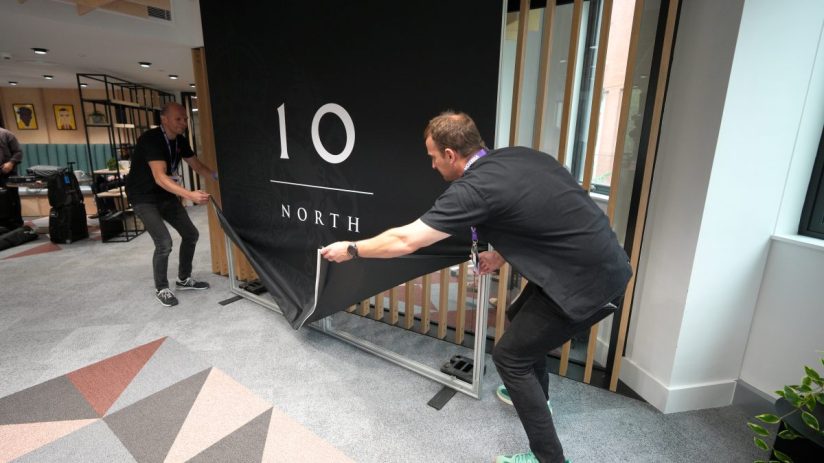Two men setting up a black banner with 10 NORTH in white text on a grey patterned carpet.