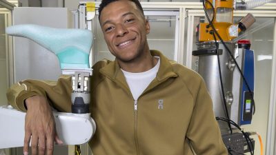 Smiling man in brown tracksuit leaning on a robotic arm with a 3D-printed shoe prototype in a lab