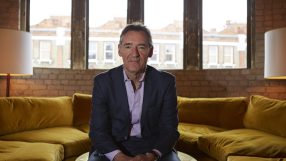 Man in suit sitting on a yellow couch in front of windows, looking directly at the camera.