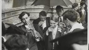 Oxford University students in academic gowns, one playing a trumpet, others drinking under an umbrella.