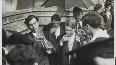 Oxford University students in academic gowns, one playing a trumpet, others drinking under an umbrella.