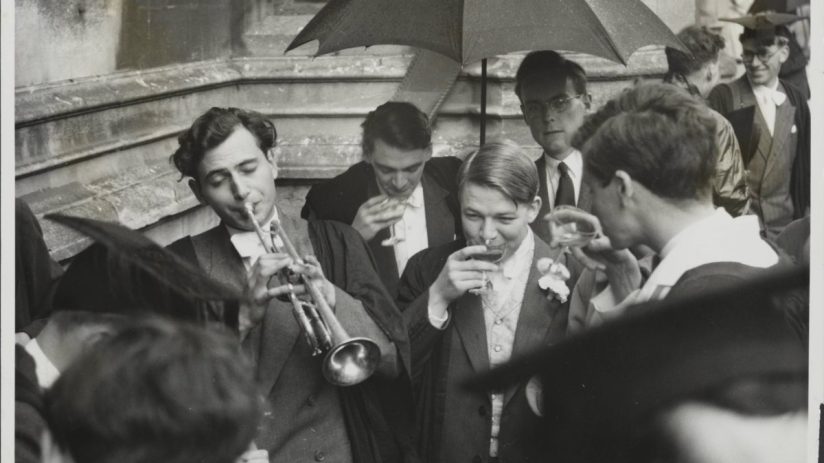 Oxford University students in academic gowns, one playing a trumpet, others drinking under an umbrella.