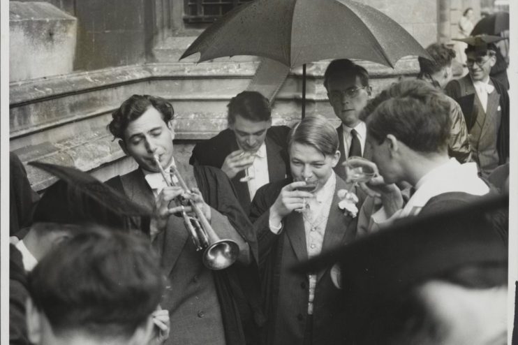 Oxford University students in academic gowns, one playing a trumpet, others drinking under an umbrella.