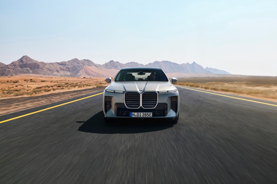 Front view of a silver BMW i7 driving on an open road through a desert landscape with mountains.
