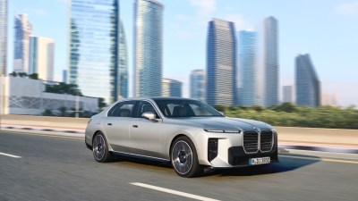 Silver 2023 BMW 7 Series sedan driving on a city road with modern skyscrapers in the background