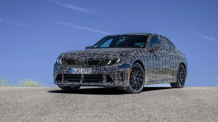Camouflaged BMW sedan prototype with black and white pattern on an asphalt road under a clear blue sky