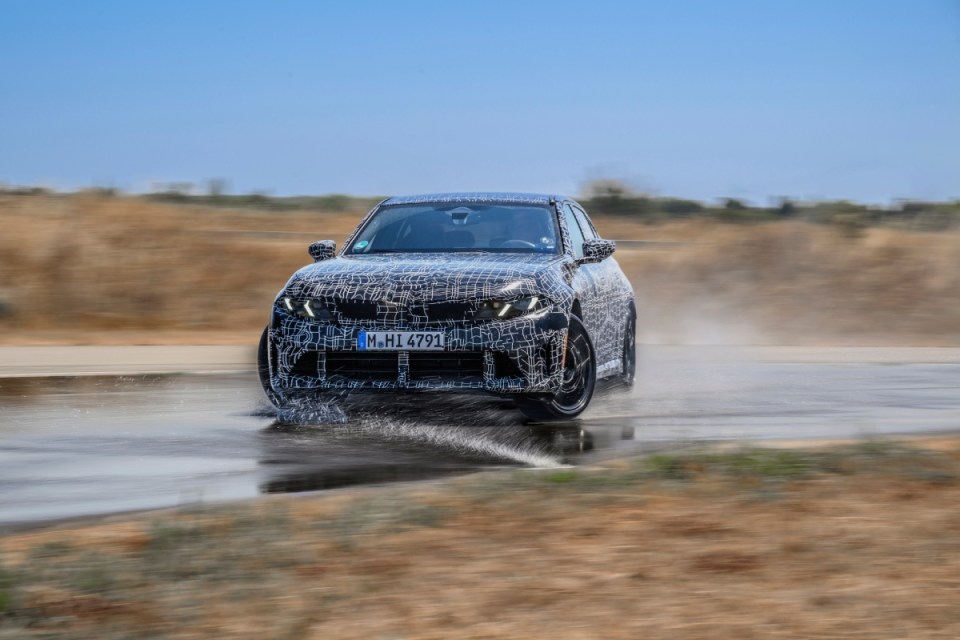 Camouflaged prototype car splashing through water on a test track, dynamic vehicle testing.