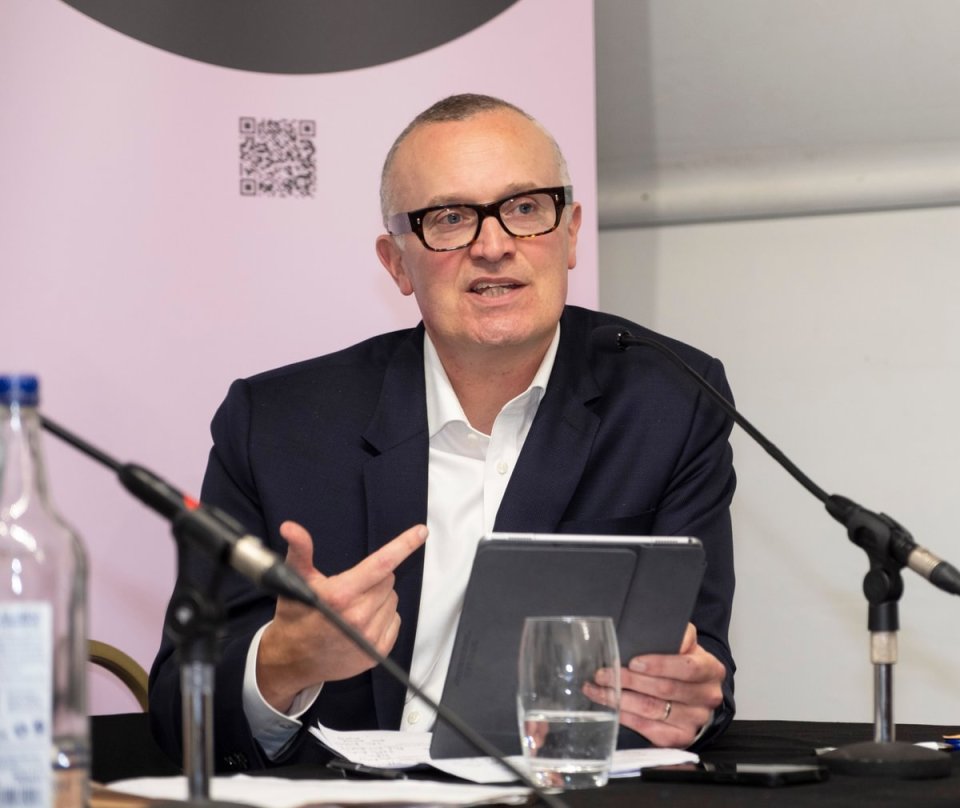Man in glasses speaking at a podium with microphones, holding a tablet at the Labour conference 2025
