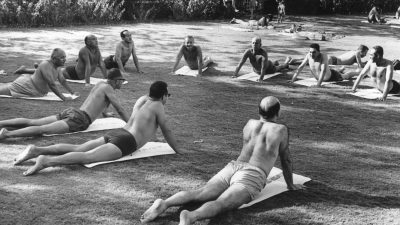 Men in swim trunks practicing yoga in a park, performing cobra pose on mats.