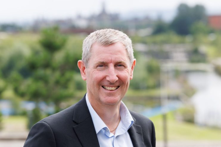 Smiling man in a suit jacket and light blue shirt, with a blurred cityscape and greenery in the background