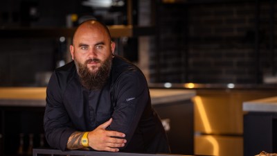Chef Nathan, a bearded man in a black chefs jacket, leaning on a railing in a modern kitchen or restaurant