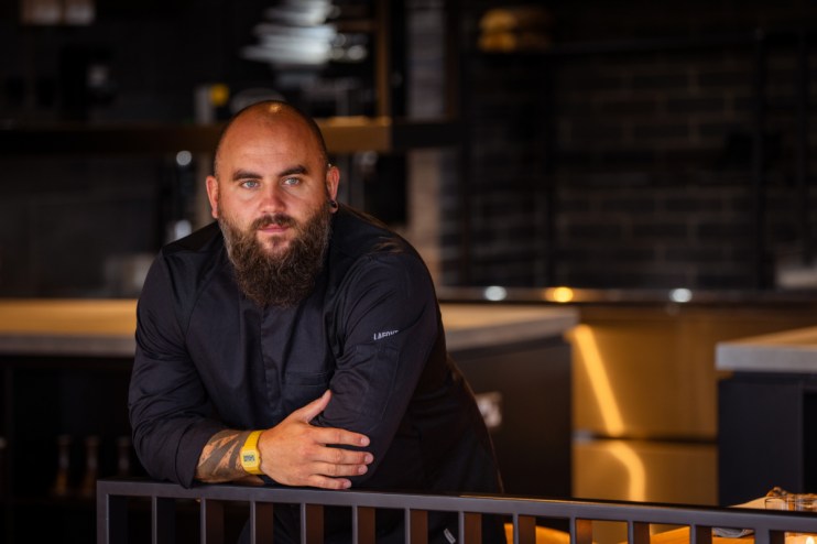 Chef Nathan, a bearded man in a black chefs jacket, leaning on a railing in a modern kitchen or restaurant