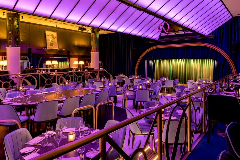 Quaglinos restaurant interior with purple lighting, set tables, and a stage with gold and green curtains.