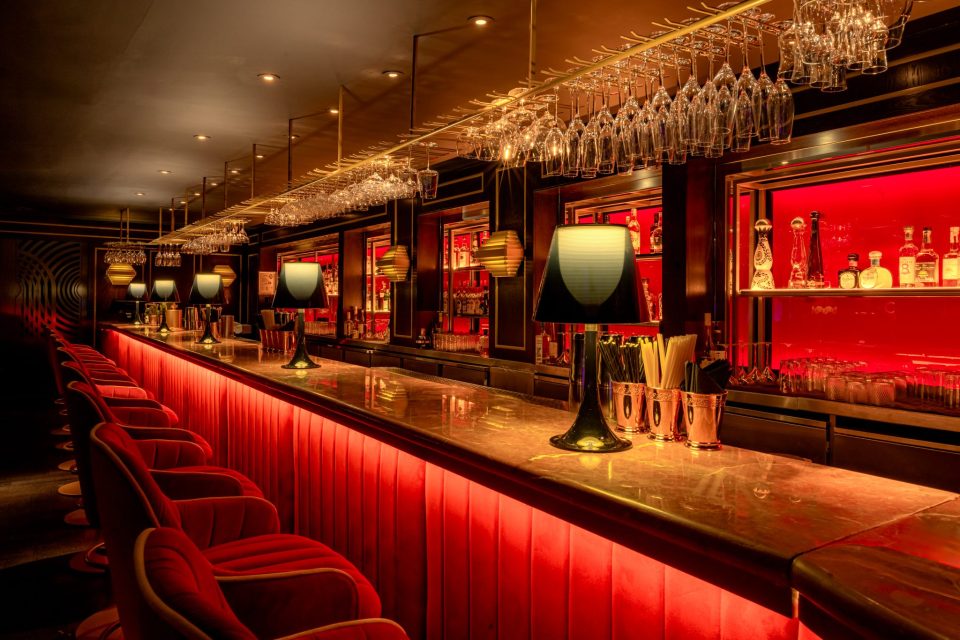 Luxurious bar interior with red velvet stools, marble counter, and illuminated shelves of bottles, Quaglinos final three.