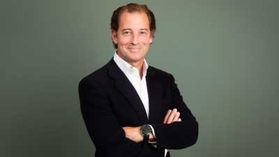 Professional man in dark blazer and white shirt, arms crossed, smiling at camera against gray background