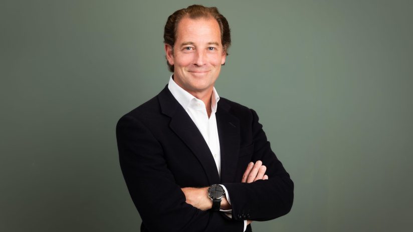 Professional man in dark blazer and white shirt, arms crossed, smiling at camera against gray background