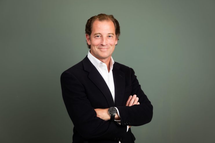 Professional man in dark blazer and white shirt, arms crossed, smiling at camera against gray background