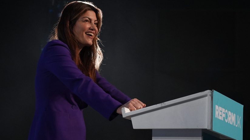 Laila Cunningham of Reform UK smiling while standing at a podium with the partys logo, speaking at an event