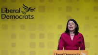 Sarah Olney speaking at a Liberal Democrats podium with party logo on a yellow backdrop