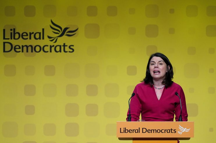 Sarah Olney speaking at a Liberal Democrats podium with party logo on a yellow backdrop