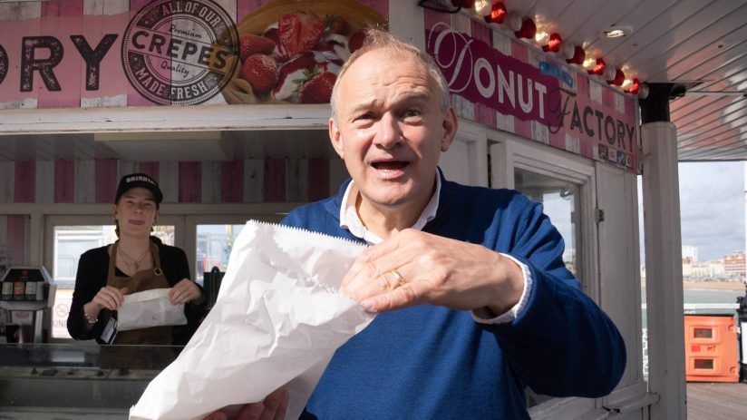 Sir Ed Davey opening a white paper bag outside a Doughnut Factory stall with a vendor in the background.