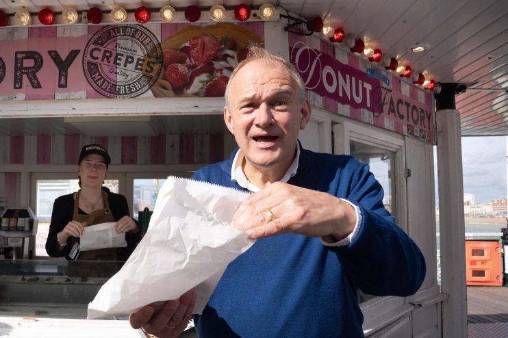 Sir Ed Davey opening a white paper bag outside a Doughnut Factory stall with a vendor in the background.