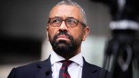 Sir James Cleverly, UK politician, wearing glasses, a suit, and a red and black patterned tie, looking upward.