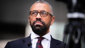 Sir James Cleverly, UK politician, wearing glasses, a suit, and a red and black patterned tie, looking upward.