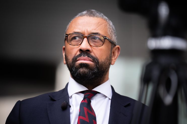 Sir James Cleverly, UK politician, wearing glasses, a suit, and a red and black patterned tie, looking upward.