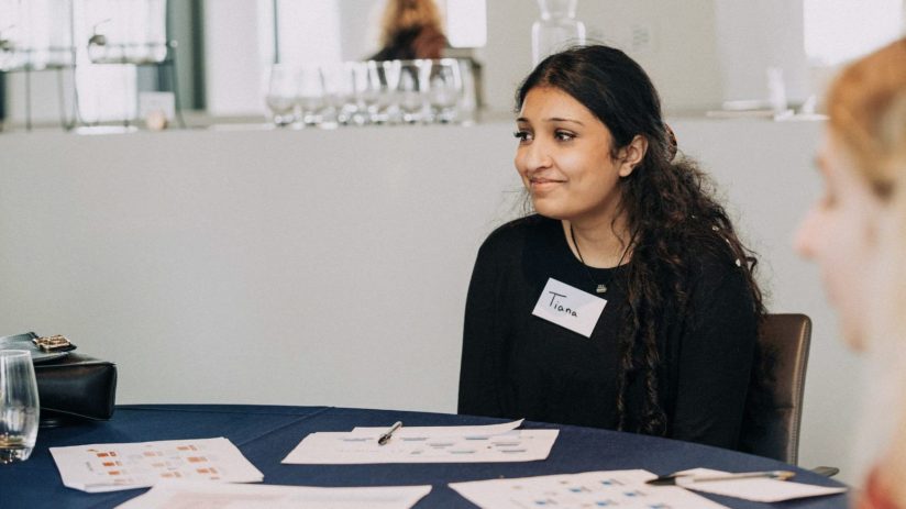 Tiana, a woman with dark hair and a name tag, smiles while seated at a table with documents for a business meeting.