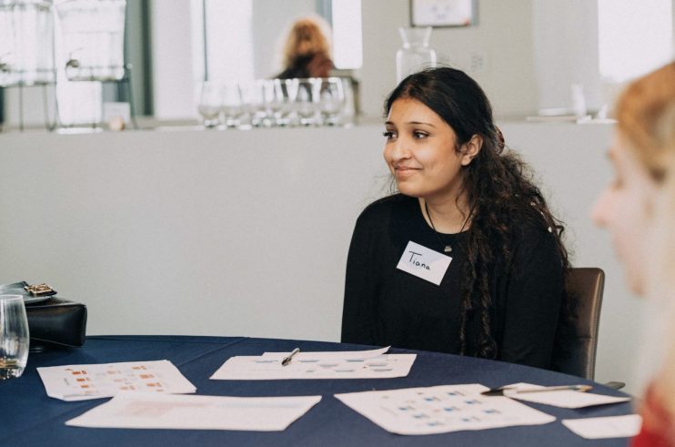 Tiana, a woman with dark hair and a name tag, smiles while seated at a table with documents for a business meeting.