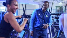 Man in The Gym Group uniform smiling at a woman on an exercise bike.