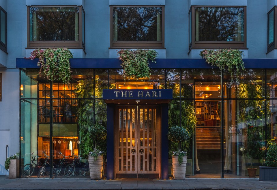 The Hari London hotel exterior at dusk, with THE HARI sign above wooden doors, glass facade, and lush greenery.