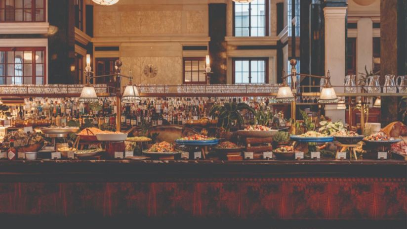 Lavish buffet spread at The Ned with diverse dishes, fresh salads, and a full bar in a grand, historic interior.