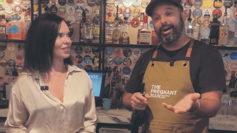 Bartender in The Pregnant Man apron speaking to a woman in a bar, bottles and coasters behind them.