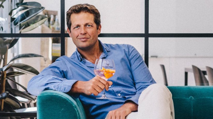 Tim Warrillow seated on a teal sofa, holding a drink with orange slices, in a modern office.