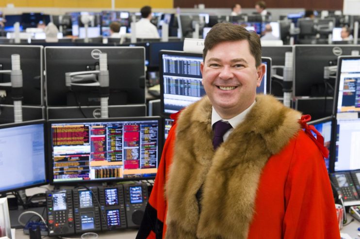 Tim Hailes smiling in official robes in a bustling trading office with multiple computer screens.