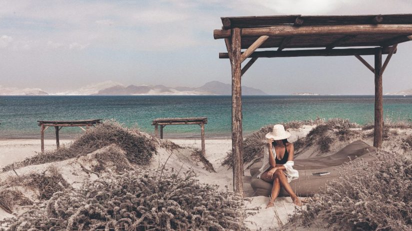 Woman in a straw hat and swimwear relaxing under a wooden pergola on a sandy beach in Bodrum, Turkey.