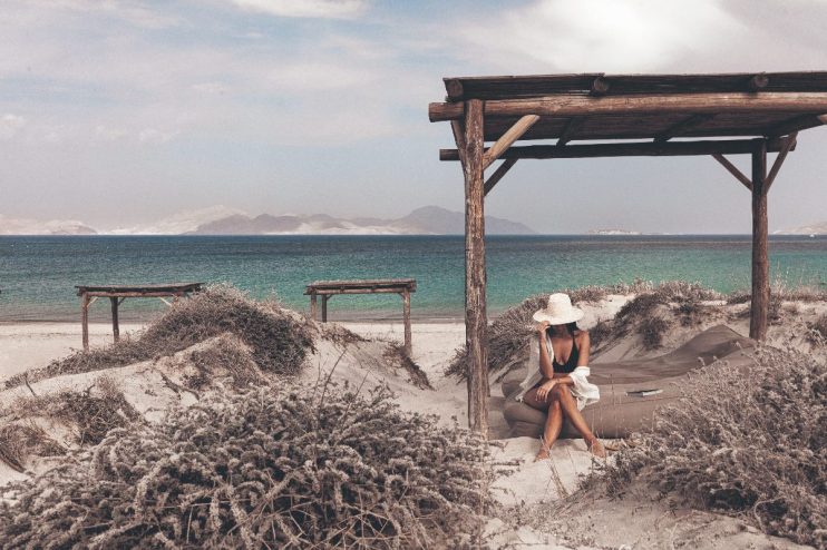 Woman in a straw hat and swimwear relaxing under a wooden pergola on a sandy beach in Bodrum, Turkey.