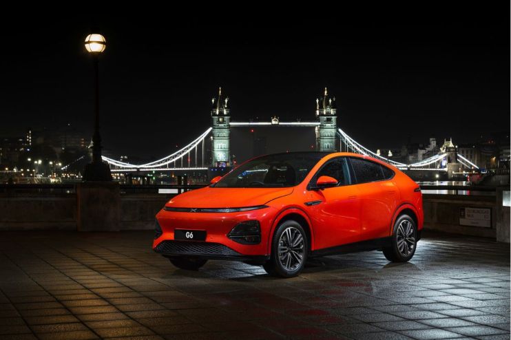 Bright orange XPeng G6 electric SUV parked on a paved street at night, with Londons Tower Bridge illuminated in the backgr...