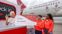 Richard Branson with Team GB athletes holding a London to Los Angeles boarding pass by a Virgin Atlantic plane.