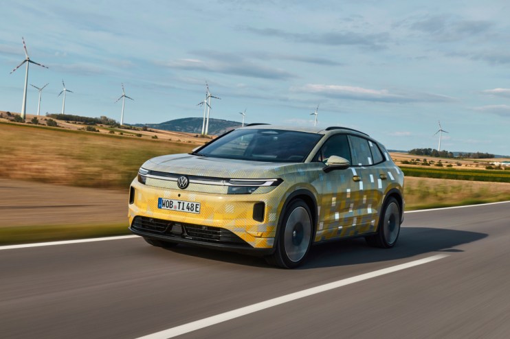 VW ID.4 Tiguan prototype in camouflage on a road with wind turbines in the background