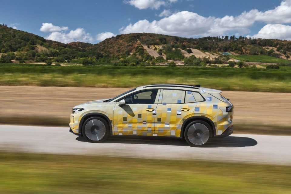 Side view of a camouflaged VW ID.4 or Tiguan prototype driving on a road, with hills and fields in the background.