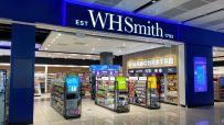 WHSmith store interior with prominent blue signage, display racks of books, travel items, and Manchester souvenirs.
