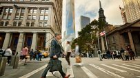 Businessman crossing a Wall Street intersection with One World Trade Center and Trinity Church in the background.