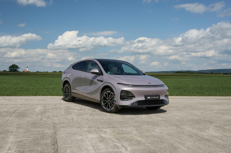 XPENG New G6 electric SUV in light purple, parked on concrete with green fields and a blue sky in the background.