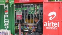 Man using phone at an Airtel Money agent booth, with M-Pesa signs and mobile phones displayed.