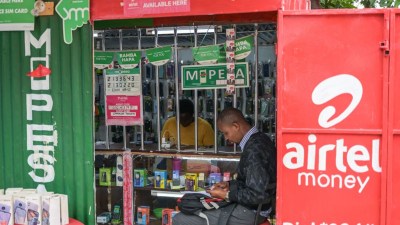 Man using phone at an Airtel Money agent booth, with M-Pesa signs and mobile phones displayed.