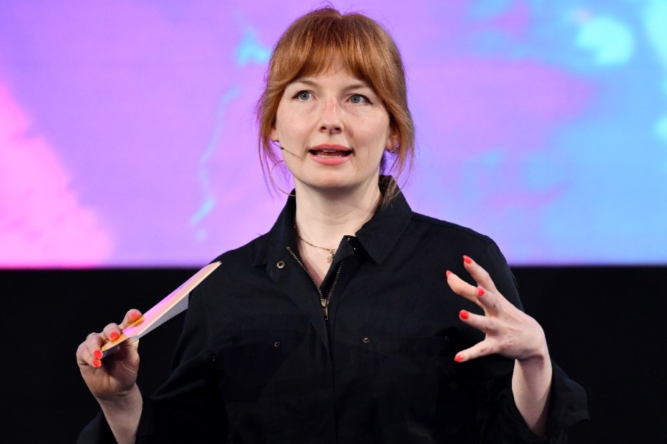 Alice Levine speaking at an event, wearing a black top and holding a colorful card, with a vibrant background