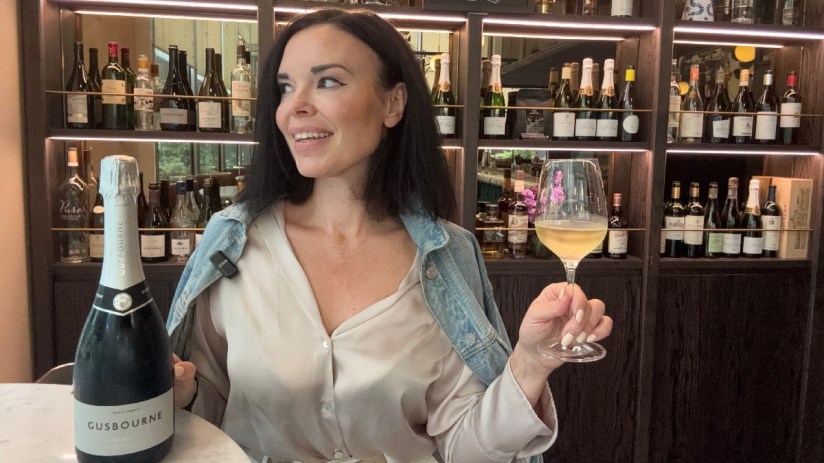 Woman smiling, holding a wine glass and standing next to a Gusbourne sparkling wine bottle at Daves Wine Bar.